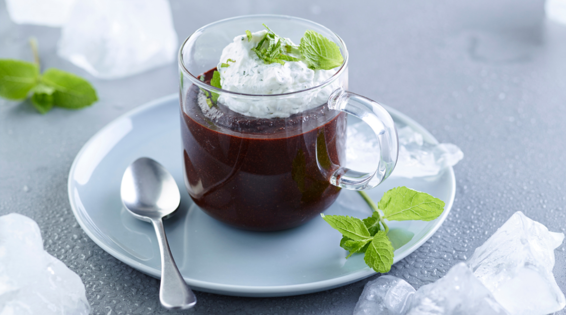 una tazza trasparente su piattino azzurro con cucchiaino di metallo contiene una granita al cioccolato con panna alla menta. Come decorazione delle foglioline di menta
