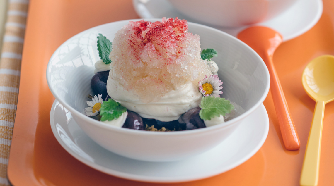 Un piatto fondo bianco su piatto piano è apparecchiato su una tavola arancione con due cucchiai colorati. Al suo interno una granita al sambuco cioccolato e ciliegie