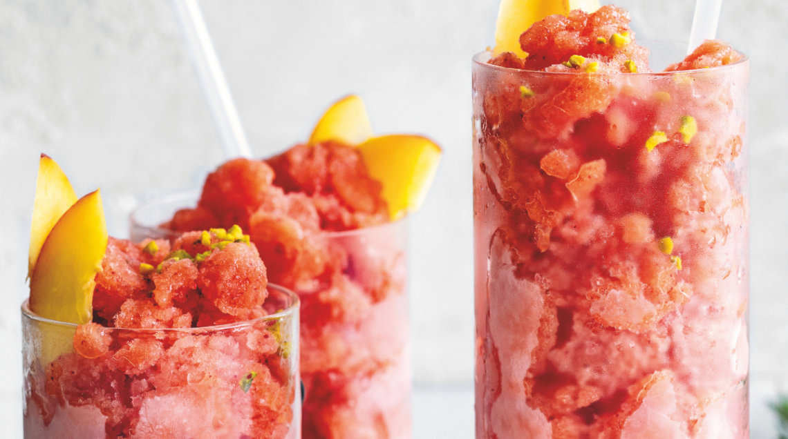 tre bicchieri alti e stretti di vetro contengono granita di pesche, rosmarino e pistacchi e delle cannucce bianche