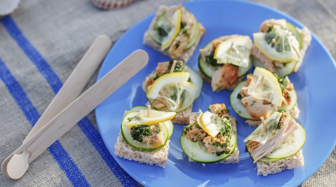 Mini tramezzini al salmone su piatto fondo blu con affianco una pinza di legno per servire gli ospiti a Ferragosto