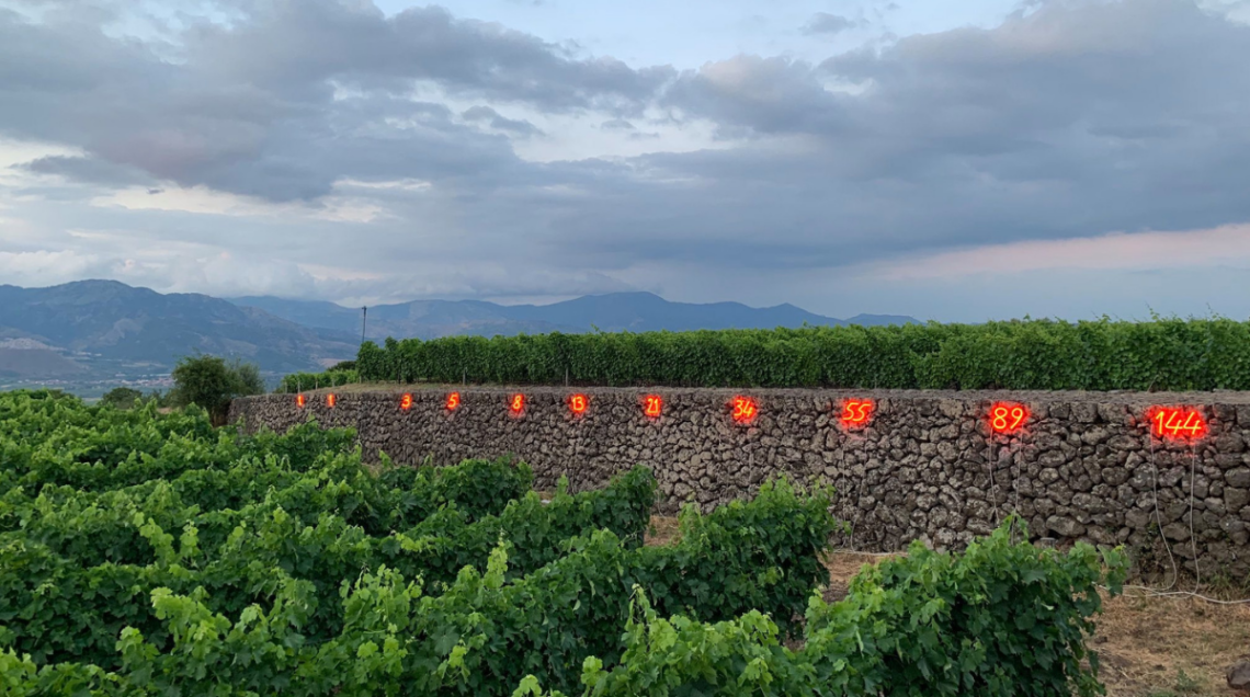 Inquadratura orizzontale ravvicinata della successione numerica a neon di Mario Merz sul muretto di pietra lavica sul vulcano.