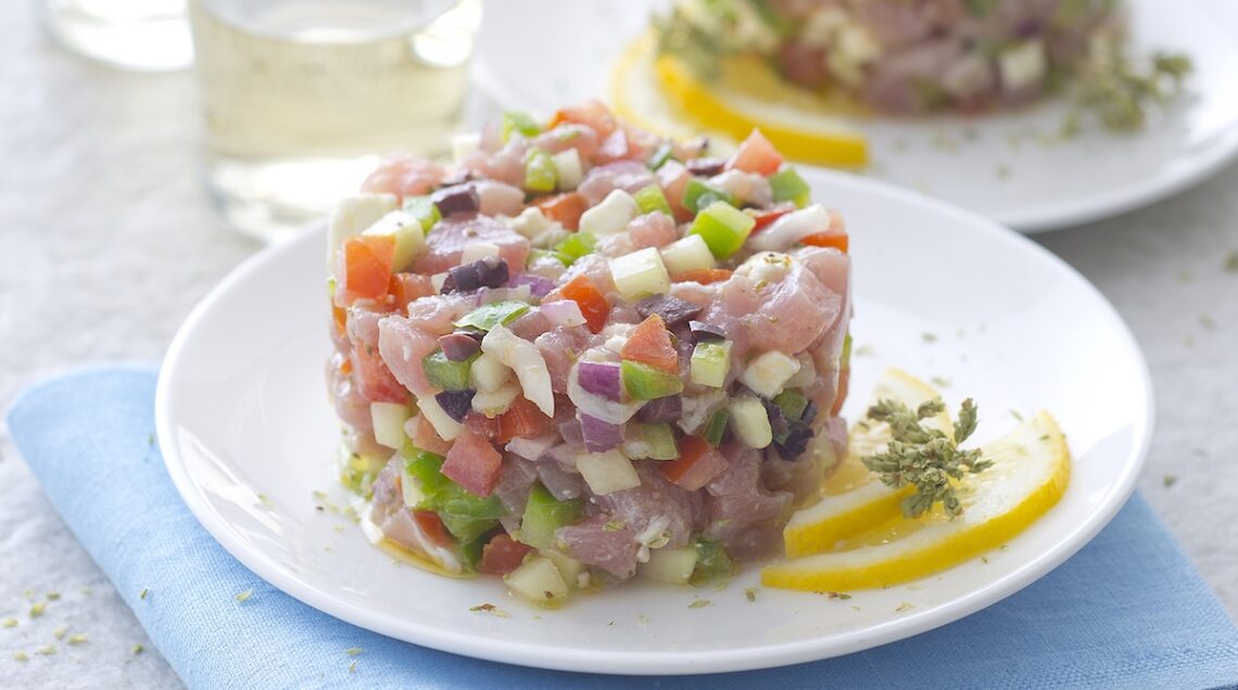 Tartare di tonno alla greca impiattata su un piatto fondo bianco. Sotto un tovagliolo azzurro