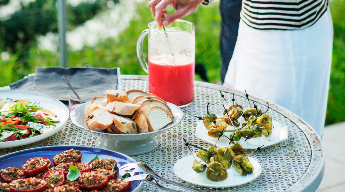 tavola di un terrazzo con diverse preparazioni per l'aperitivo e una mano che gira un cocktail rosso in una caraffa