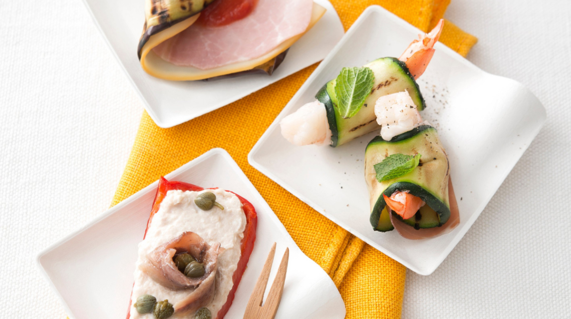 Tre piattini bianchi quadrati con finger food colorati. Un peperone con crema e acciuga, rotolini di melanzana con prosciutto e involtini di zucchina con gamberi e menta