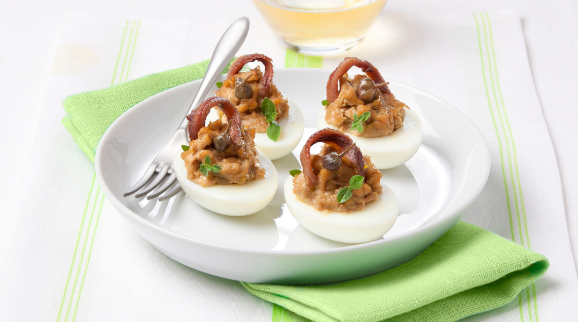 Un piatto fondo bianco contiene quattro uova ripiene con mousse e vitello tonnato, decorate con acciughe e una forchetta di metallo. Sotto il piatto un tovagliolo verde e dietro un bicchiere