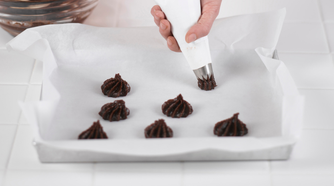 Mano che usa una sac-&agrave;-poche con punta a stella per modellare baci al cioccolato su una teglia con carta forno