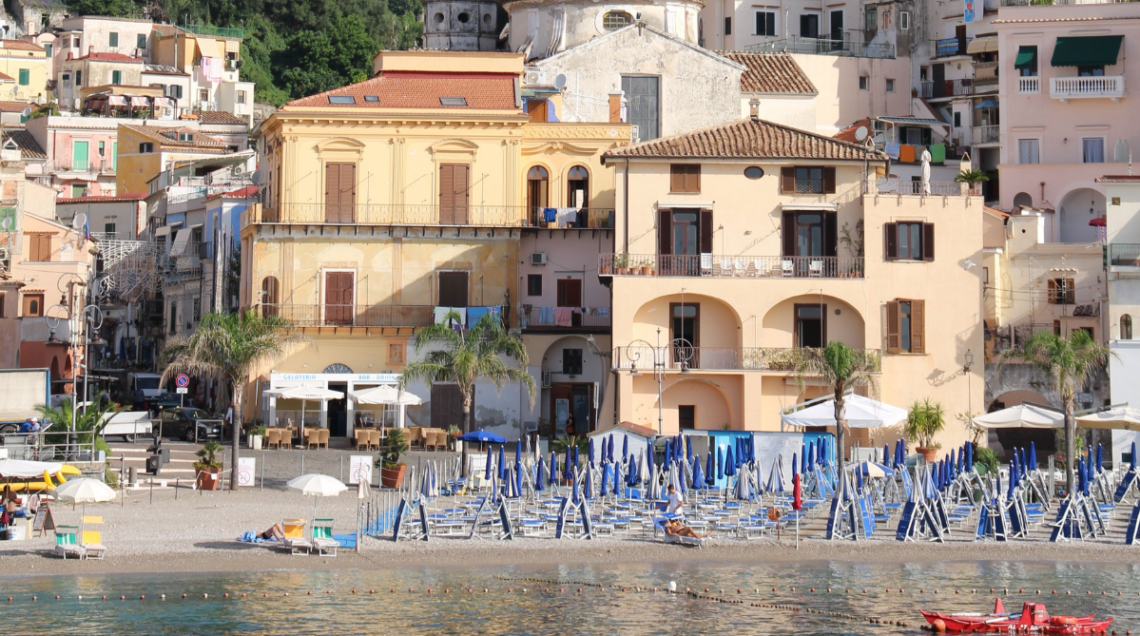 Scorcio del borgo marinaro di Cetara, in Costiera Amalfitana, ripreso dal mare. In primo piano la spiaggia con stabilimento balneare caratterizzato da file di ombrelloni blu e lettini; sullo sfondo le tipiche case colorate e gli edifici storici arroccati sulla collina.