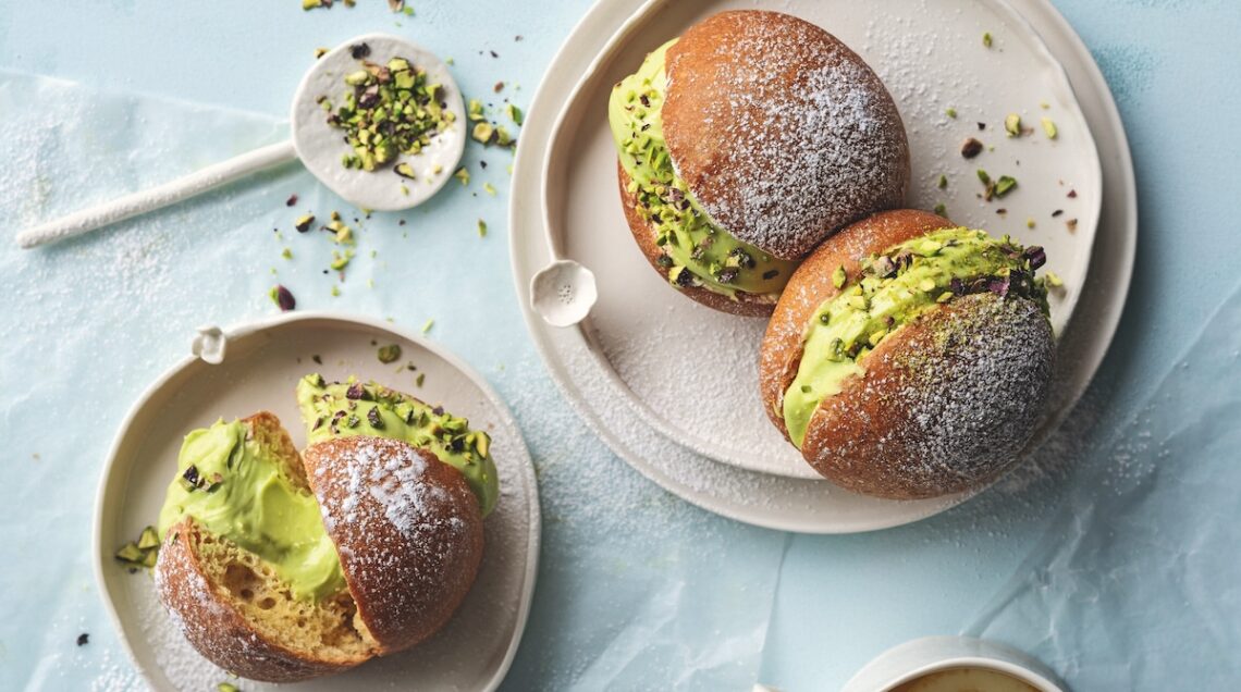 brioche con crema al pistacchio: una su un piatto piccolo a sinistra e due su un piatto tondo grande a destra. I piatti sono bianchi e la tovaglia &egrave; azzurro chiaro