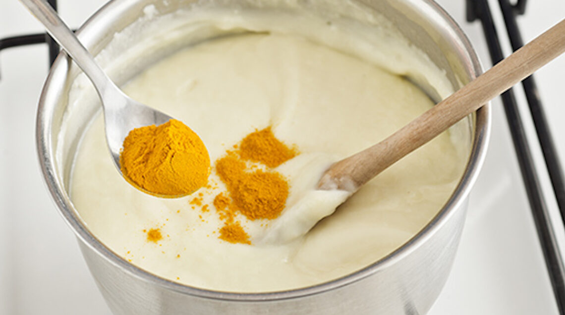 Un cucchiaino di curcuma in polvere viene versato all'interno di una salsa calda messa sul fuoco. Nella pentola è presente anche un cucchiaino di legno