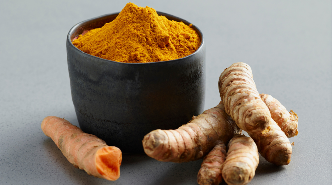 Una ciotola scura colma di curcuma in polvere circondata da alcune radici fresche della stessa spezia