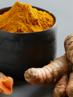 Una ciotola scura colma di curcuma in polvere circondata da alcune radici fresche della stessa spezia