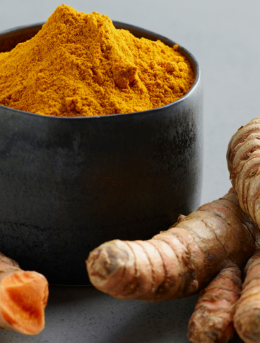Una ciotola scura colma di curcuma in polvere circondata da alcune radici fresche della stessa spezia