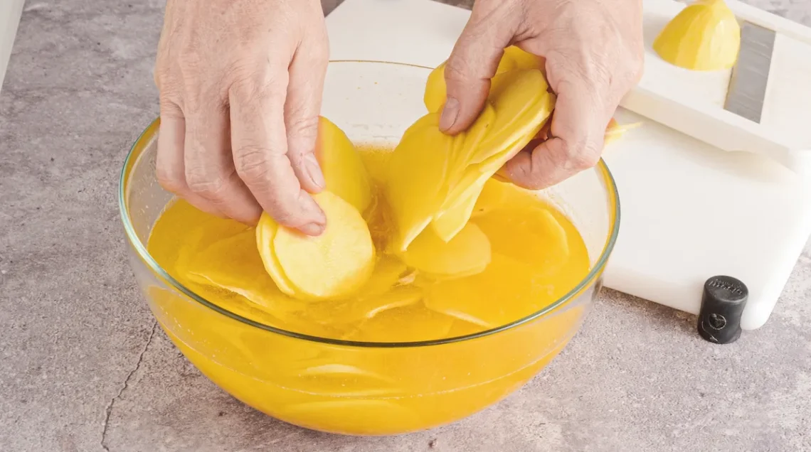 Due mani poggiano delicatamente delle fettine sottili di patate in una ciotola trasparente piena di acqua e curcuma