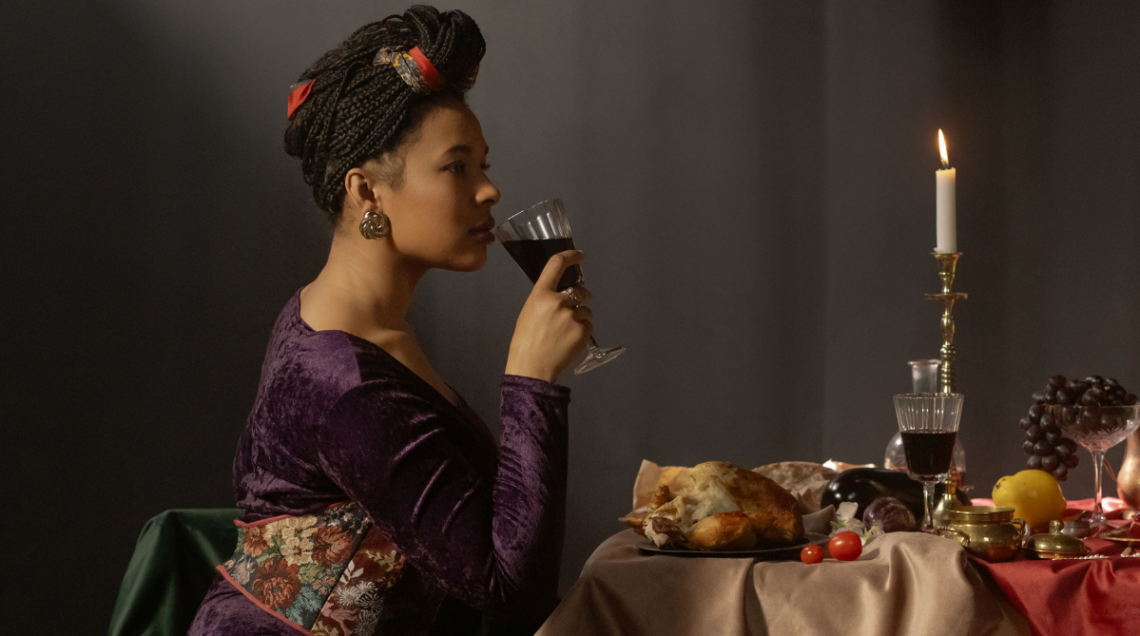 Una donna elegante di profilo sorseggia del vino rosso a una tavola imbandita con un pollo arrosto, frutta e una candela accesa in un'atmosfera classica