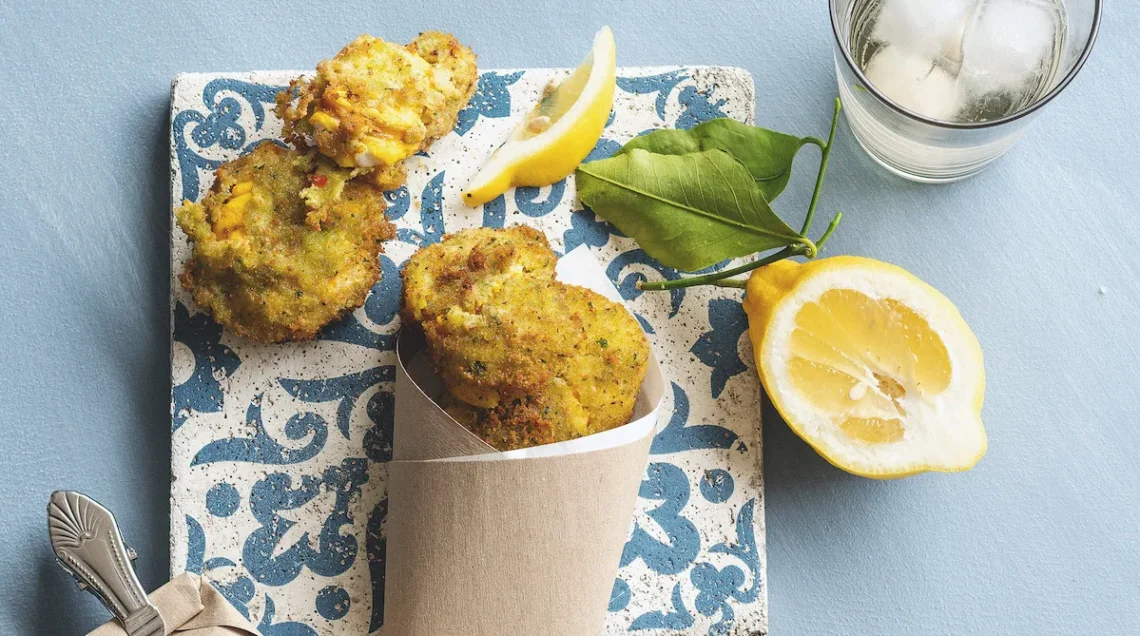 Frittini dorati e croccanti serviti in un cono di carta e distribuiti su una mattonella decorata in blu e bianco, accompagnati da mezzo limone fresco con foglie e un bicchiere con ghiaccio su uno sfondo azzurro