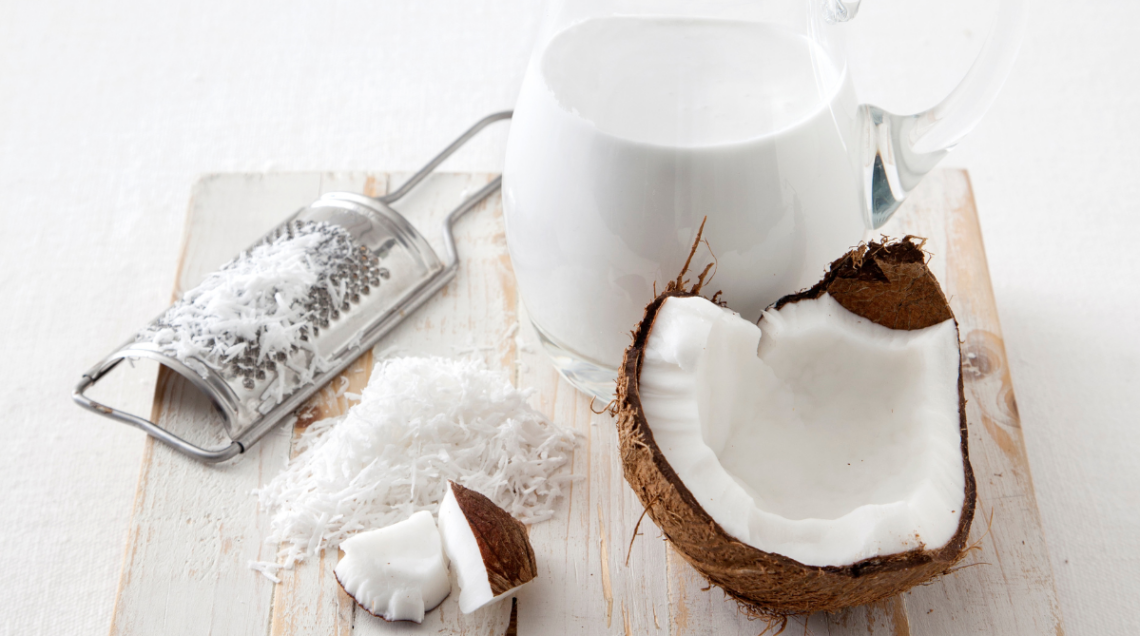 Sopra un tagliere di legno ci sono: a sinistra una grattugia, sotto del cocco grattuggiato, affianco un pezzo di cocco e sopra una brocca di vetro trasparente con del latte di cocco