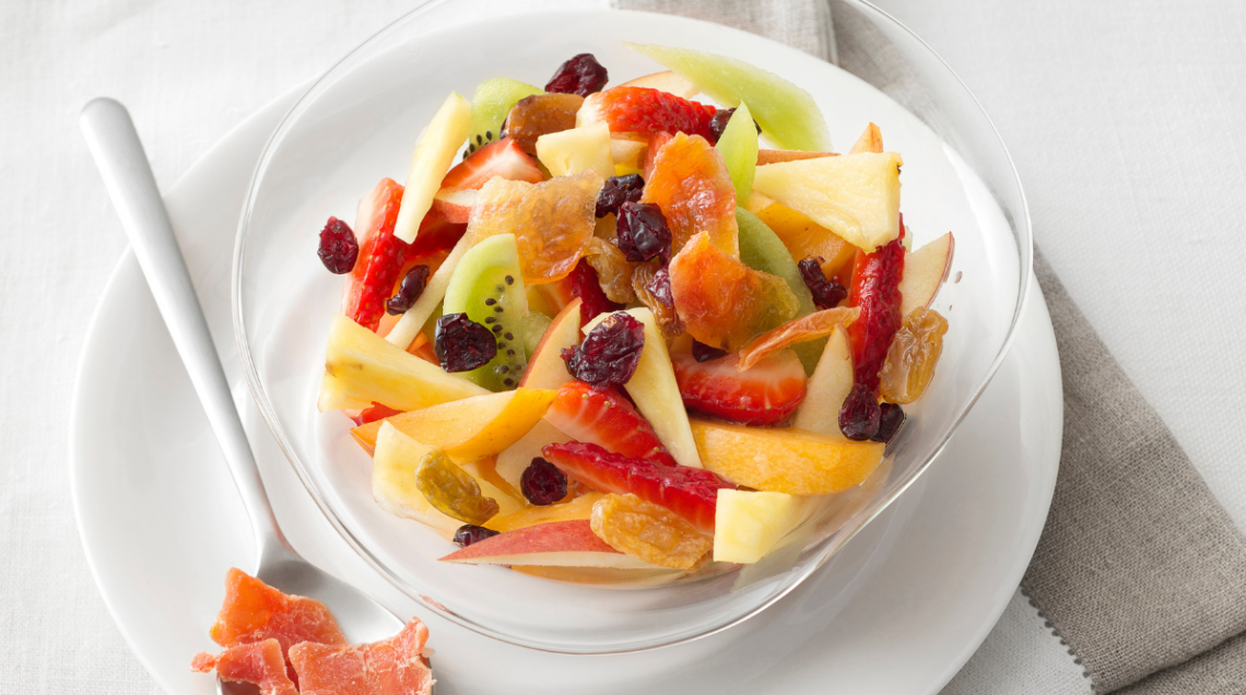una ciotola di macedonia con frutta fresca e frutta secca su piatto bianco, con cucchiaio di metallo