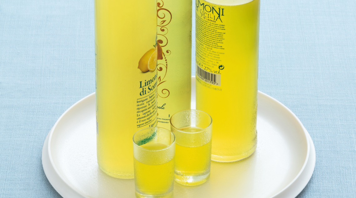 Tre bottiglie di limoncello di Sorrento, tipico liquore della Costiera Amalfitana, con due bicchierini di vetro ripieni. Il tutto su un piatto rotondo bianco.