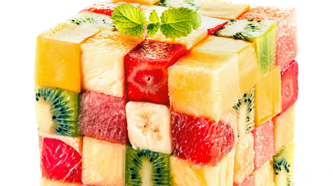 Un cubo geometrico ispirato al cubo di Rubik, formato da cubetti perfetti di frutta assortita (come kiwi, fragola, banana, anguria e melone) e sormontato da una fogliolina di menta su sfondo bianco.