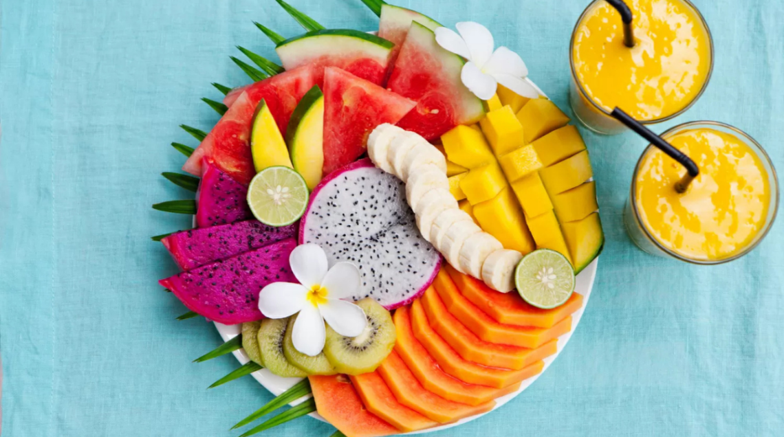 Un piatto rotondo visto dall'alto ricco di frutta tropicale affettata con cura (mango, papaya, kiwi, dragon fruit e anguria), decorato con fiori bianchi e affiancato da due frullati gialli su sfondo azzurro