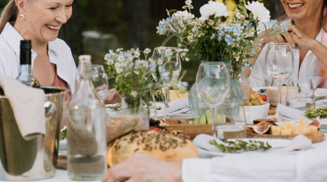 Momento conviviale all'aperto attorno a una tavola imbandita con pane, formaggi, calici di vino e vasi ricchi di fiori di campo. Nello sfondo due donne sorridono
