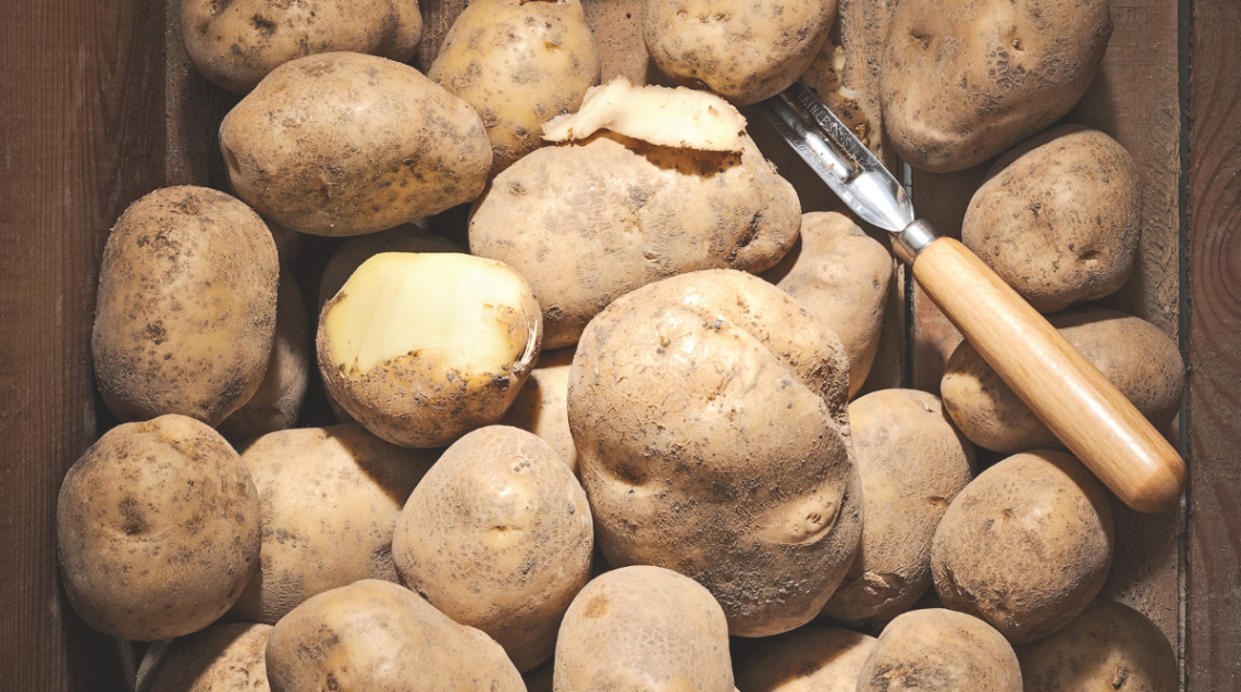 Una cassetta di legno piena di patate terrose. Una di queste è stata parzialmente pelata e c'è un pelapatate con manico in legno a destra.