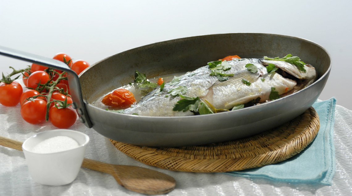 Pesce all'acqua pazza in padella sopra un tagliere di legno. Vicino dei pomodorini, un piccolino contenitore aperto con del sale fino e un cucchiaio di legno. La cucina tipica della Costiera Amalfitana