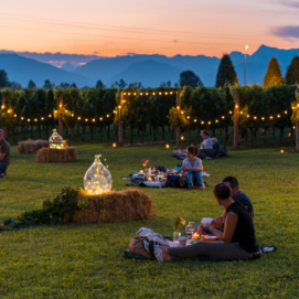 Persone che fanno un picnic serale in un vigneto illuminato da file di luci calde al tramonto, con montagne sullo sfondo.