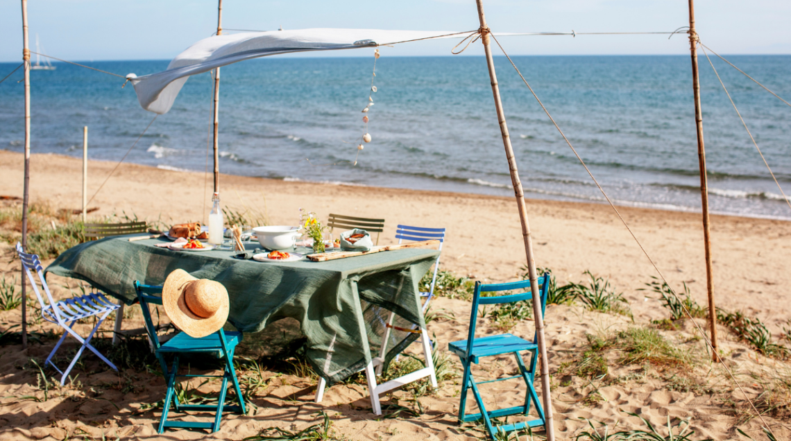 tavola imbandita per il pranzo in spiaggia fresco e leggero. Intorno, sedie pieghevoli colorate, un cappello di paglia appeso e il mare blu sullo sfondo.