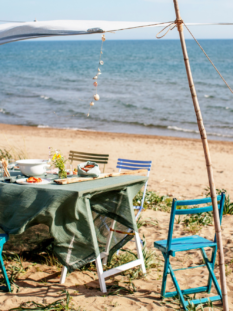 tavola imbandita per il pranzo in spiaggia fresco e leggero. Intorno, sedie pieghevoli colorate, un cappello di paglia appeso e il mare blu sullo sfondo.