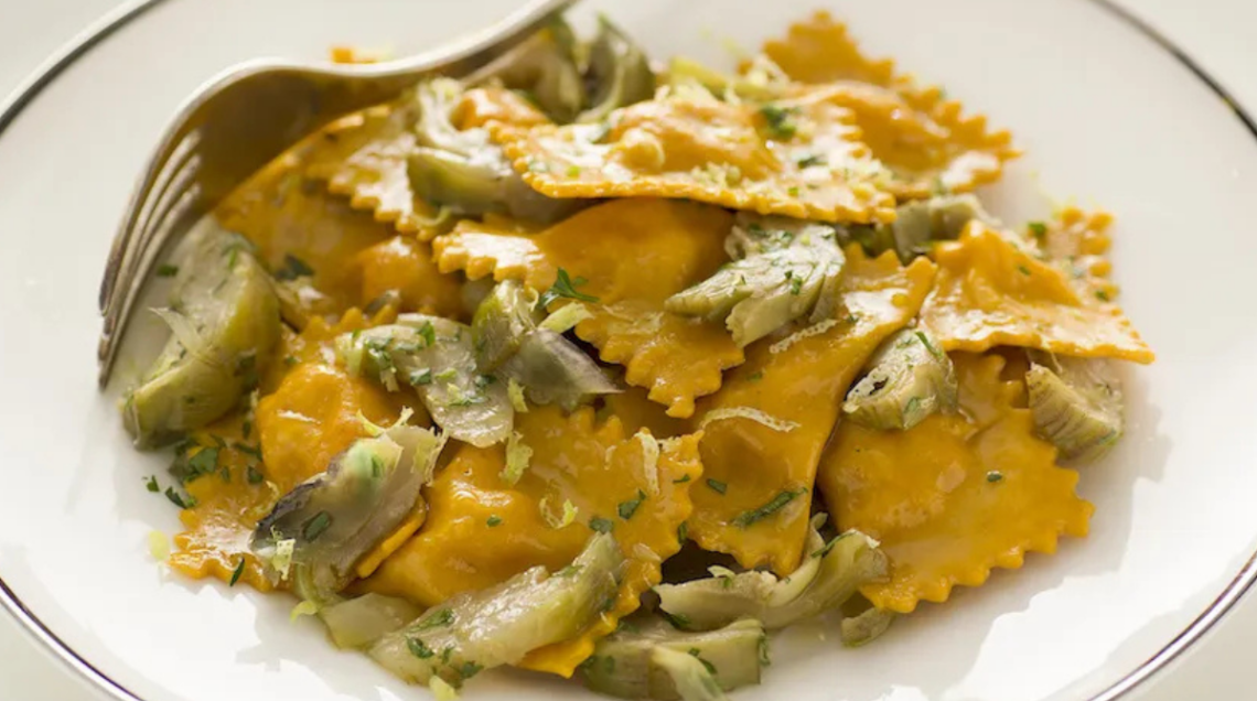 Un piatto fondo bianco con forchetta di metallo contiene dei ravioli di scampi con curcuma e salsa ai carciofi