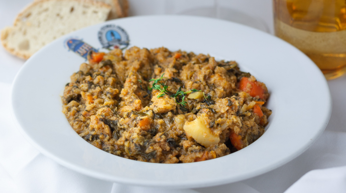 Ribollita in un piatto bianco. Sullo sfondo un pezzo di pane e un bicchiere arancione