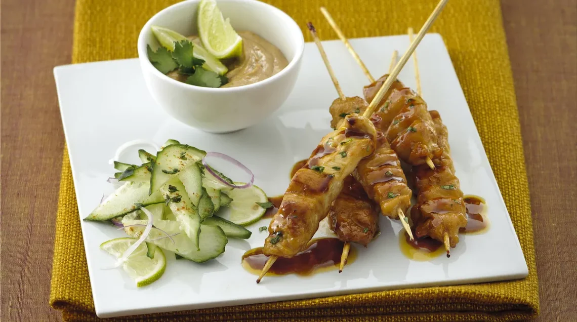 Un piatto quadrato bianco contiene dei bastoncini di satay alla curcuma,