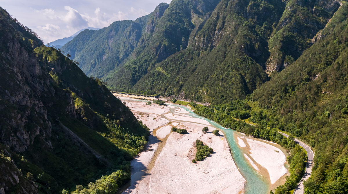 Veduta aerea della valle del Tagliamento in Friuli-Venezia Giulia, che mostra un vasto letto di ghiaia bianca e un canale di acqua turchese cristallina che serpeggia tra imponenti e fitte montagne boscose. Una strada costeggia il fiume sul lato destro.