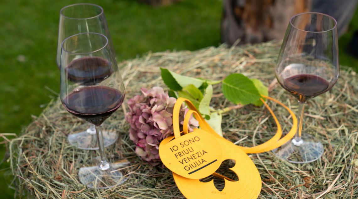 Tre calici di vino rosso su una balla di fieno con un talloncino giallo "Io sono Friuli Venezia Giulia".
