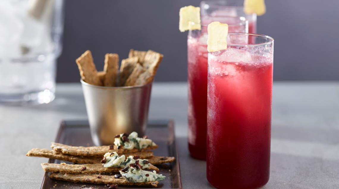 Cocktail con vino rosso all'interno di tre bicchieri alti e stretti di vetro, decorati con zenzero candito. Affianco a questi, sulla sinistra, un piattino con dei mini cracker di semi fatti in casa