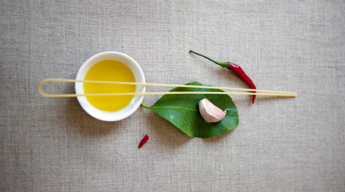 su sfondo neutro, partendo da sinistra, ci sono: una ciotolina bianca con olio, una foglia con uno spicchio d'aglio appoggiato sopra e un peperoncino. Uno spaghetto &egrave; messo sopra a tutti questi ingredienti