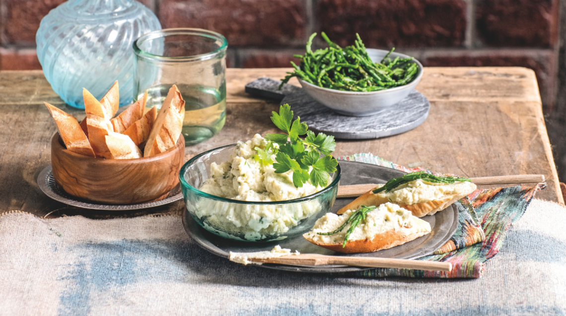 Su un tavolo di legno rustico con un muro di mattoni sullo sfondo, sono disposti una ciotola di crema spalmabile bianca con prezzemolo, due crostini con samphire (salicornia), pane tostato e bicchieri d'acqua