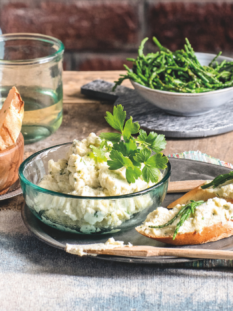 Su un tavolo di legno rustico con un muro di mattoni sullo sfondo, sono disposti una ciotola di crema spalmabile bianca con prezzemolo, due crostini con samphire (salicornia), pane tostato e bicchieri d'acqua