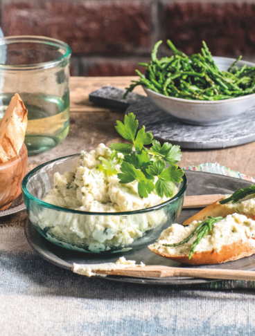 Su un tavolo di legno rustico con un muro di mattoni sullo sfondo, sono disposti una ciotola di crema spalmabile bianca con prezzemolo, due crostini con samphire (salicornia), pane tostato e bicchieri d'acqua