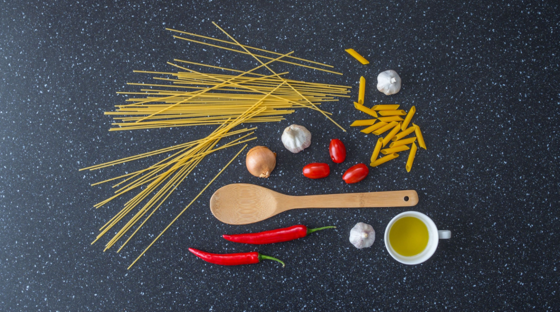 Su sfondo neutro: spaghetti, aglio, cipolla, penne, peperoncino, ciotolina bianca con olio e cucchiaio di legno