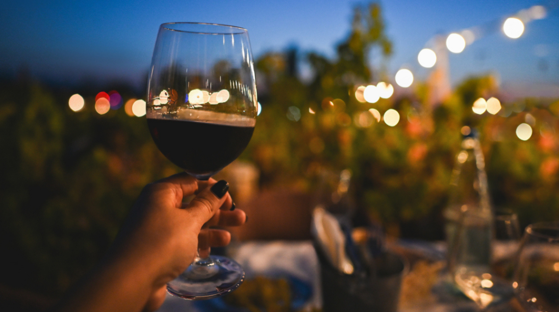 Un calice di vino rosso tenuto in mano con sfondo romantico di un giardino con lucin