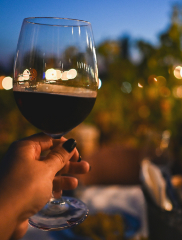 Un calice di vino rosso tenuto in mano con sfondo romantico di un giardino con lucin