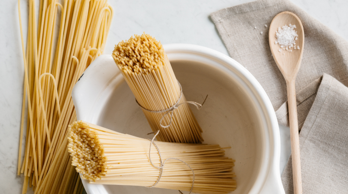 a sinistra, in verticale, degli spaghetti sfusi. Affianco una ciotola bianca contiene degli spaghetti chiusi con un nastrino. A destra un cucchiaio di legno con del sale grosso &egrave; posto sopra un canovaccio beige