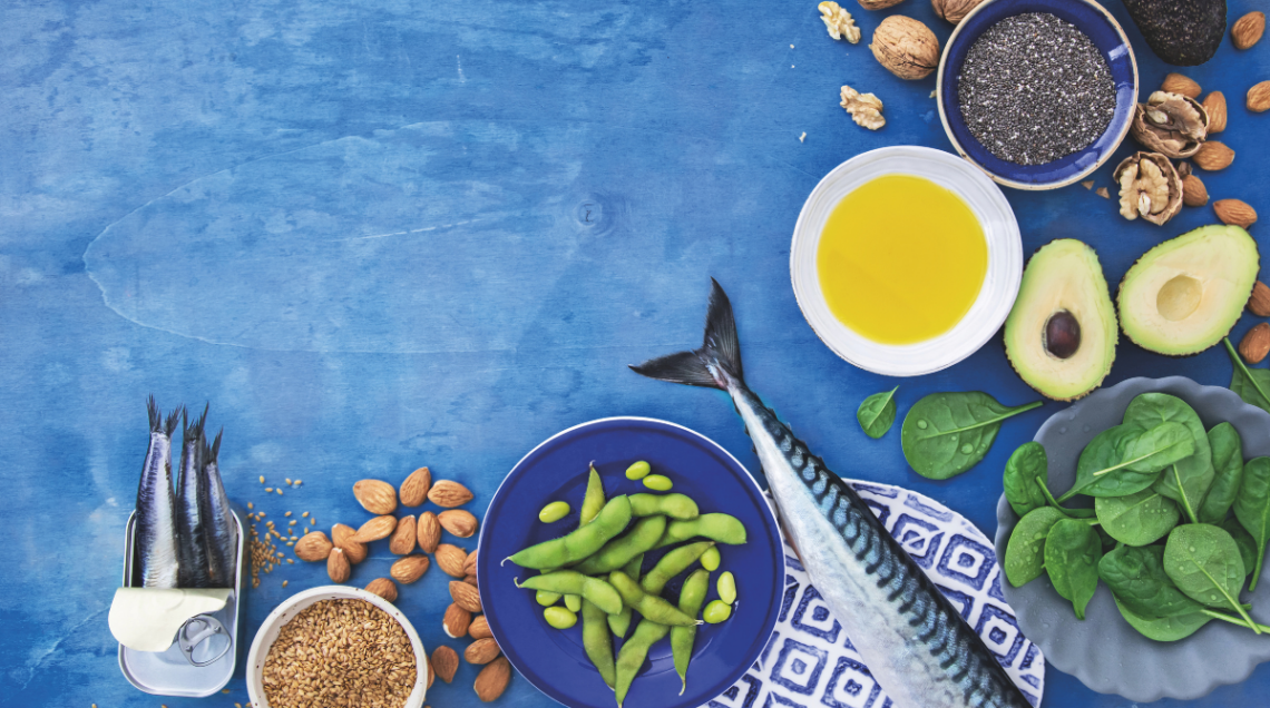 Diversi alimenti su sfondo azzurro, come le noci, l'avocado, l'olio, le mandorle, gli spinacini, il pesce azzurro