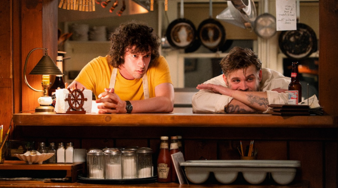 Dominic Sessa e Leo Woodall appoggiati alla cucina stanno ascoltando e guardando con attenzione qualcosa
