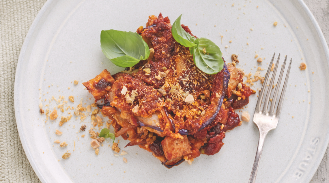 Una parmigiana di melanzane decorata con basilico fresca servita su piatto piano bianco con forchetta di metallo
