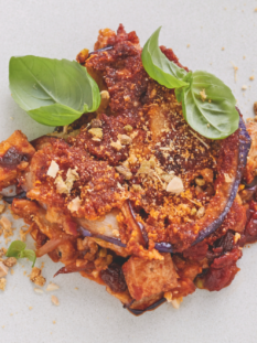 Una parmigiana di melanzane decorata con basilico fresca servita su piatto piano bianco con forchetta di metallo