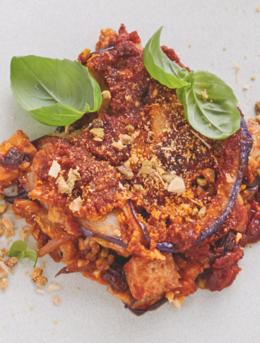 Una parmigiana di melanzane decorata con basilico fresca servita su piatto piano bianco con forchetta di metallo