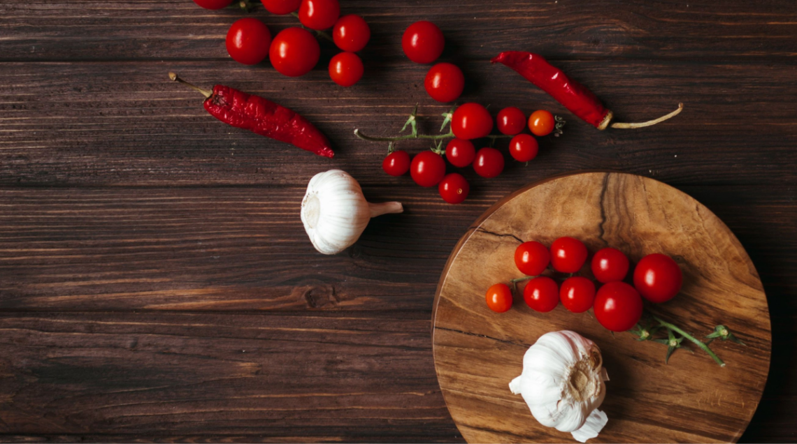 A destra in basso un tagliere rotondo di legno con pomodorini e una testa d'aglio. Gli stessi ingredienti poggiano sul tavolo di legno, insieme a due peperoncini